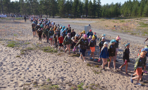 Kymmeniä partiolaisia partiopaidat päällä kävelevät hiekkarannalla jonossa.